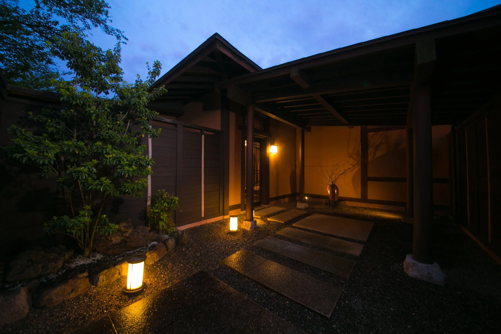 夜の日本家屋の入り口で、暖かな照明が石畳や庭木を照らし、落ち着いた雰囲気を醸し出しています。
